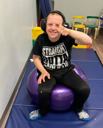 Participant with Down syndrome smiling while seated on a purple exercise ball during a fitness activity.