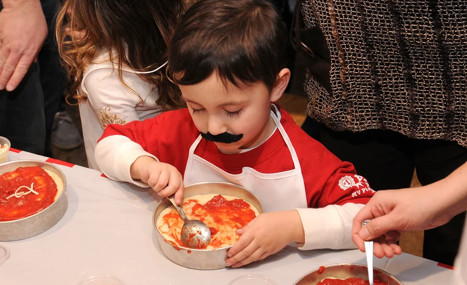Child wearing a chef apron and mustache making a personal pizza with tomato sauce.