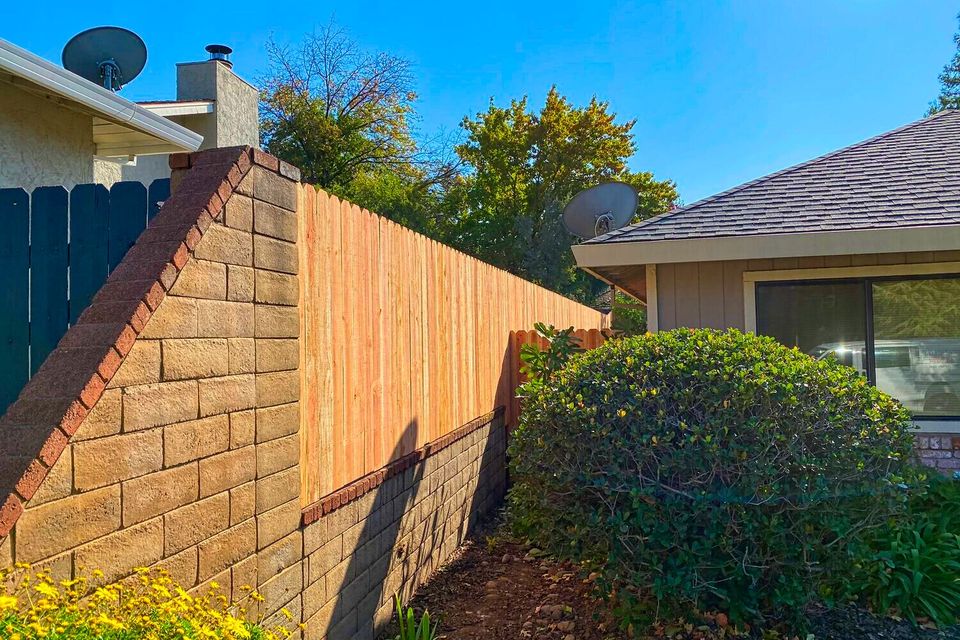 Enhanced view of a residential backyard with a new wood privacy fence atop a concrete retaining wall, bordered by yellow flowers and manicured shrubs, with neighboring homes and trees under a clear blue sky