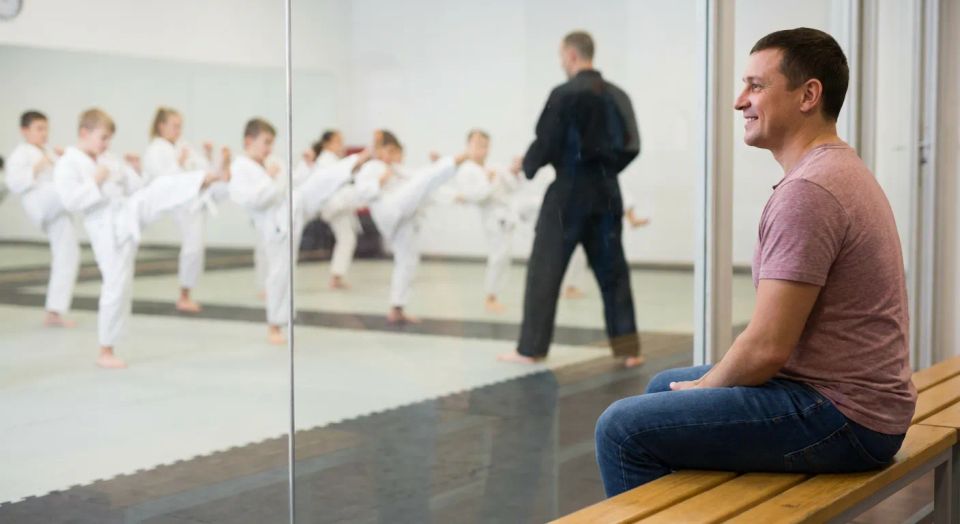Smiling father sitting on a wooden bench watching his children participate in a kids karate class through a safe glass viewing window.