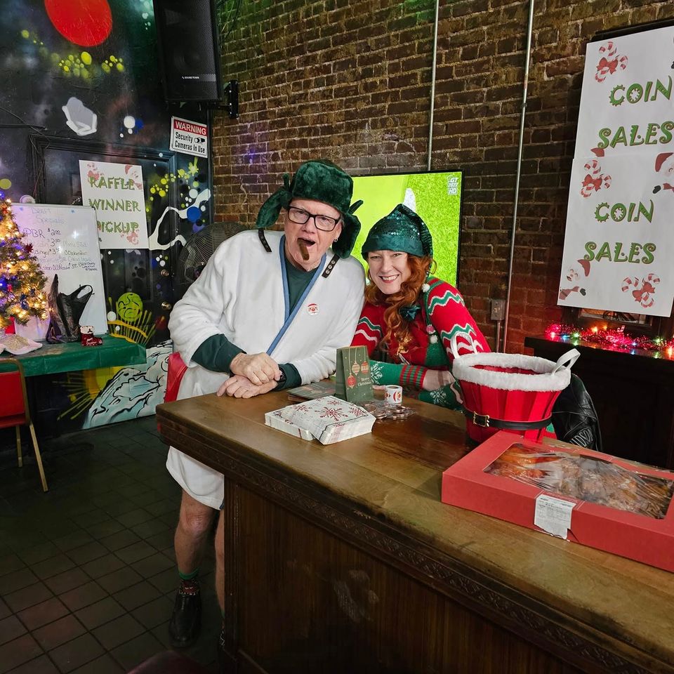 Two people in festive holiday outfits standing behind a table at a Christmas-themed fundraiser.