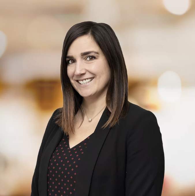 Professional portrait of Jen wearing a black blazer and patterned top, smiling against warm blurred background