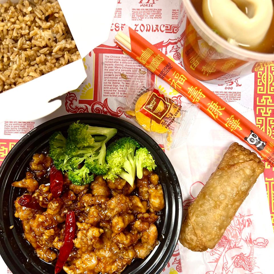 Takeout meal with entrée, fried rice, soup, egg roll, broccoli, chopsticks, and fortune cookie.