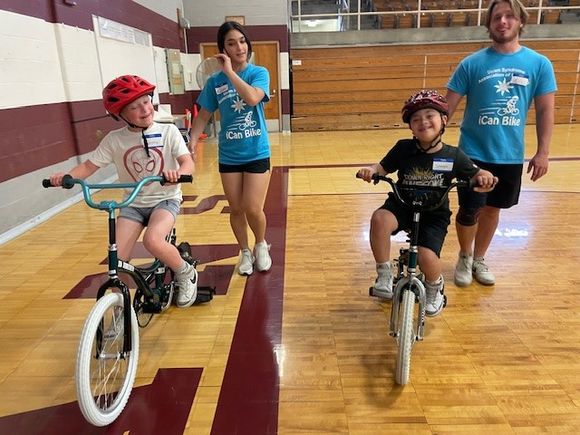 Two children ride adaptive bicycles inside a gymnasium while volunteer coaches walk beside them during DSAV's iCan Shine Bike Camp.