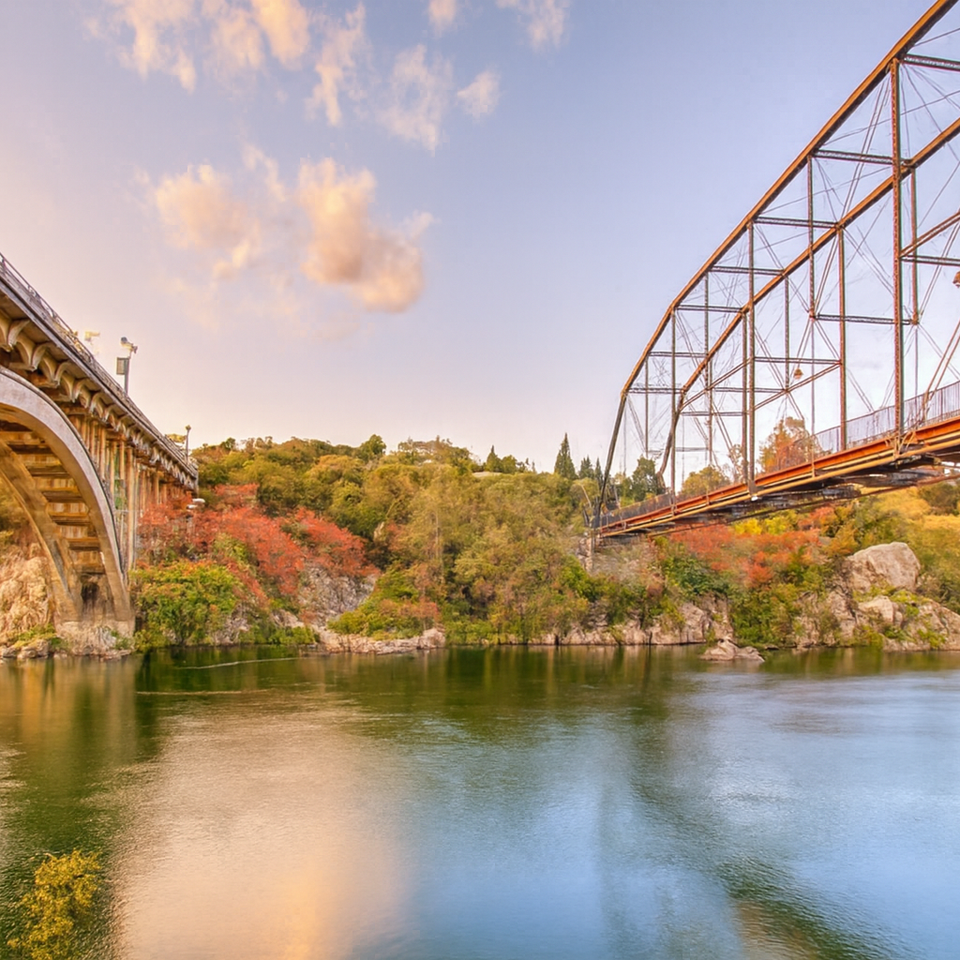 Rainbow Bridge and historic truss bridge over the American River in Folsom, CA at sunset with autumn trees and kayaker below