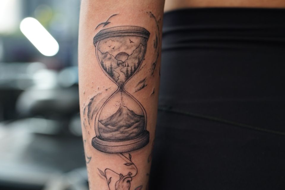 Image of an hour glass tattoo in black and grey that appears to be showing to build a reality you must first destroy a reality, excellent detail outside the hourglass as well