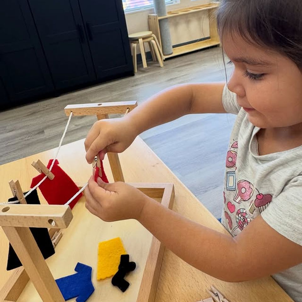 Preschool girl clipping felt clothing to a wooden clothesline during practical life work in Palmdale CA