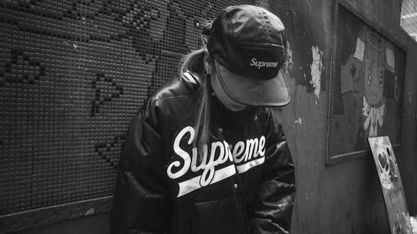 Person in a city, head tipped down with "Supreme" on their ball cap and jacket- all in black and white
