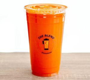 A clear cup of bright orange carrot juice with ice, labeled "The Blend Smoothies," sitting beside a whole orange carrot on a wooden table.
