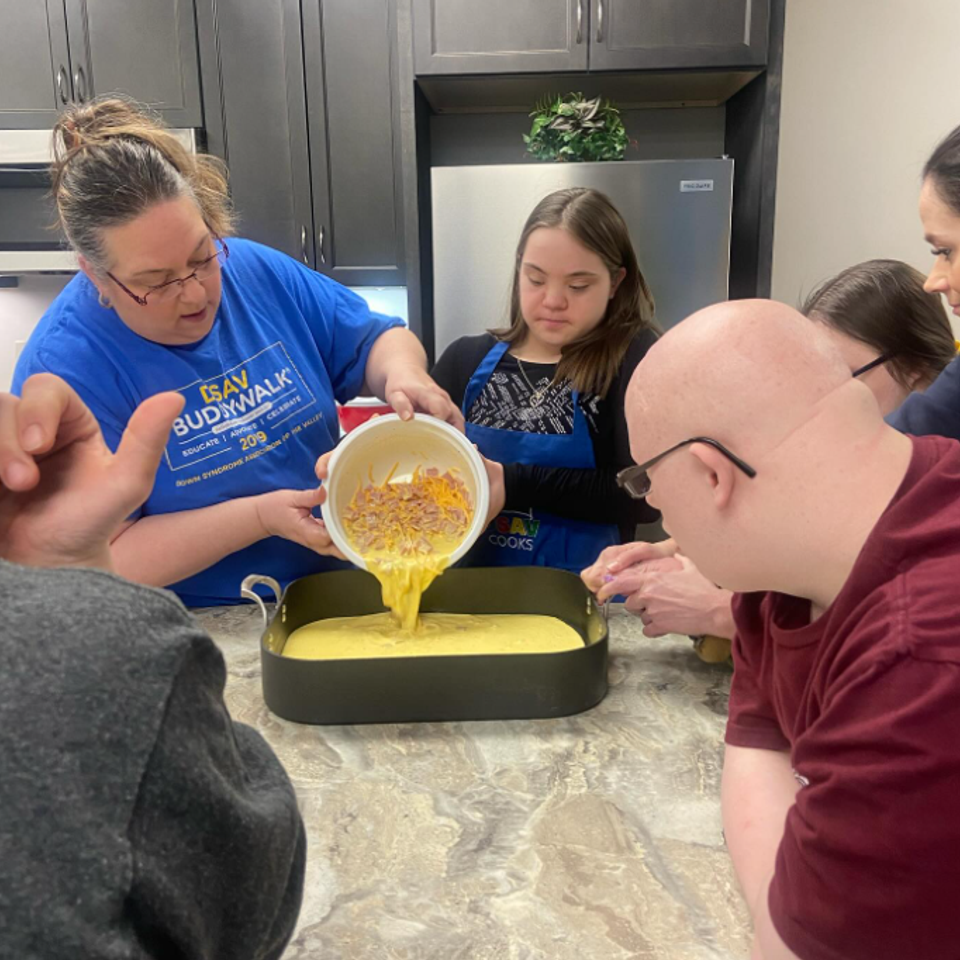 Participants with Down syndrome and instructors preparing a recipe together by pouring ingredients into a baking dish during a DSAV Cooks class.