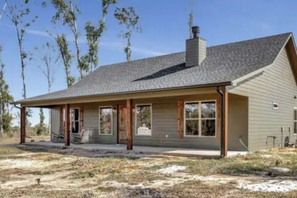 Green barndominium with covered front porch, cedar posts, and stone chimney on a rural lot