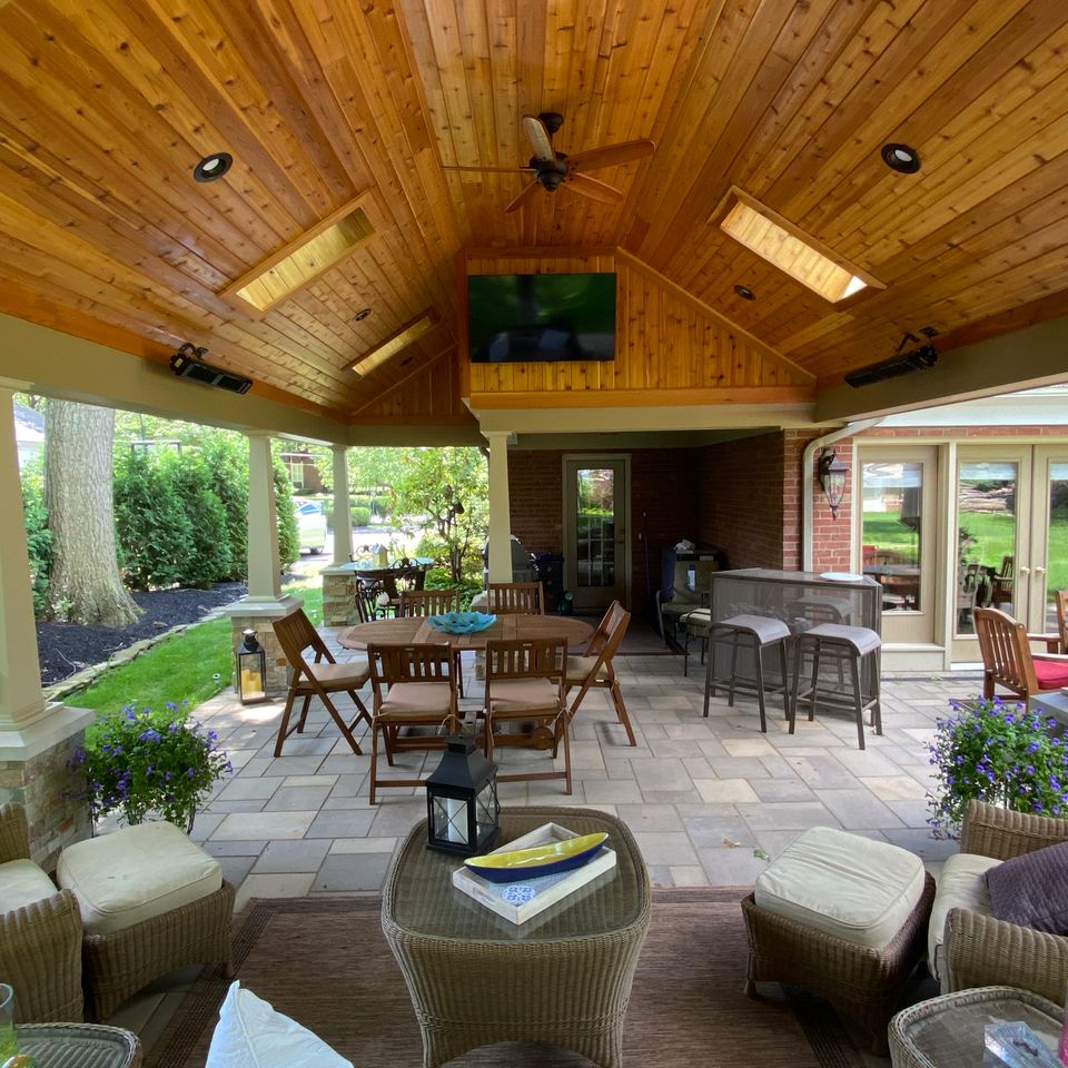 Covered outdoor living area featuring a wood-paneled ceiling, dining area, lounge seating, television, and paved patio.