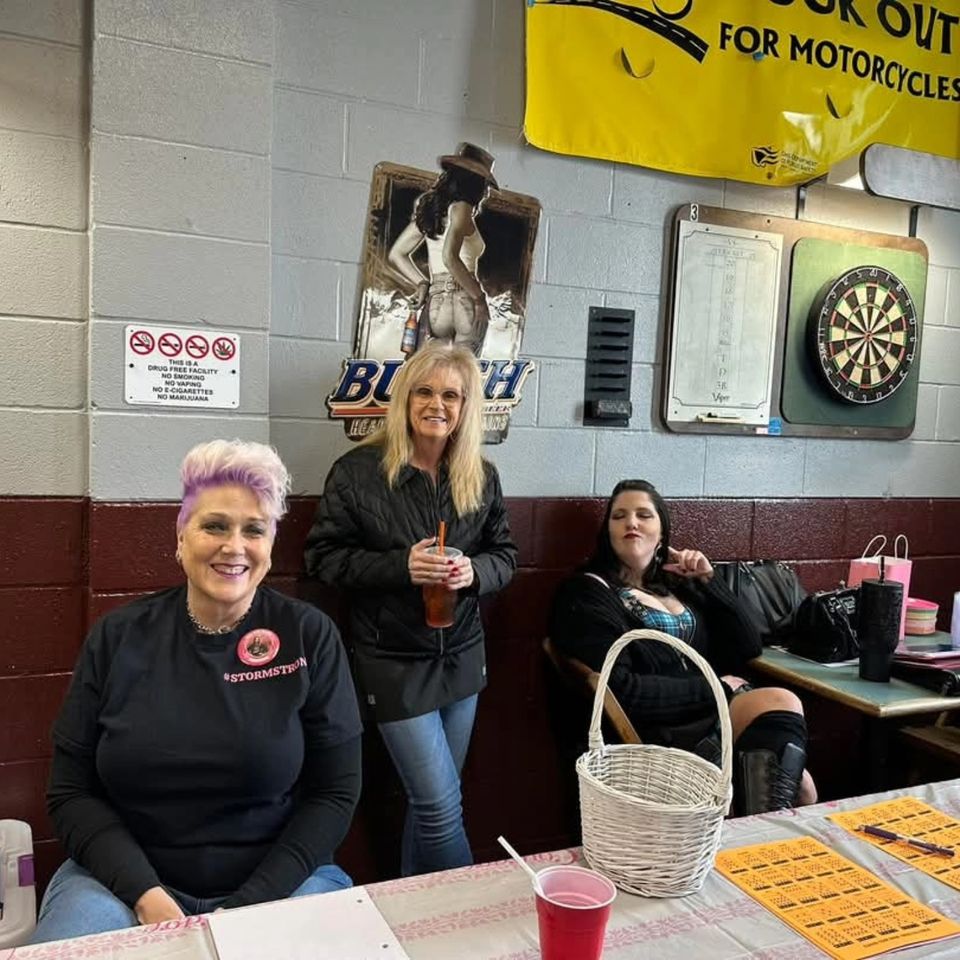 Kelly and two friends smiling at a bingo event, seated near bingo cards, raffle materials, and refreshments inside a social hall.