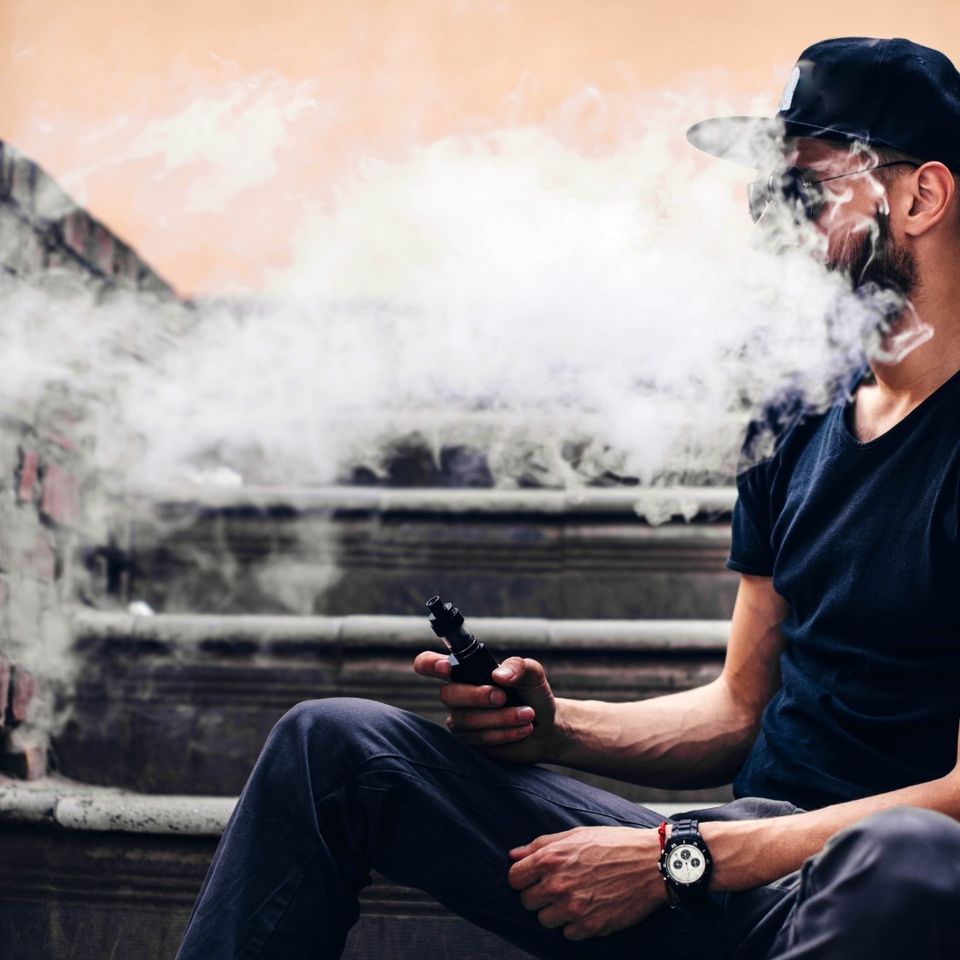 Cool man with snapback hat and aviators enjoying a vape break on a brick staircase.