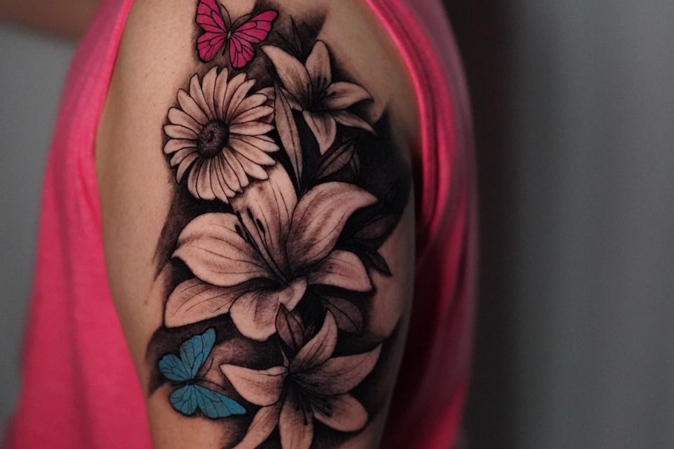 Image of the left shoulder of a man with a detailed tattoo of flowers in black & white and 2 butterflies, one is blue the other is pink