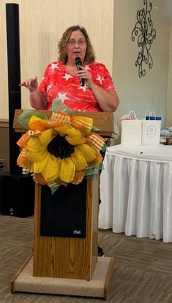 Speaker holding a microphone and addressing attendees from a podium decorated with a sunflower arrangement.