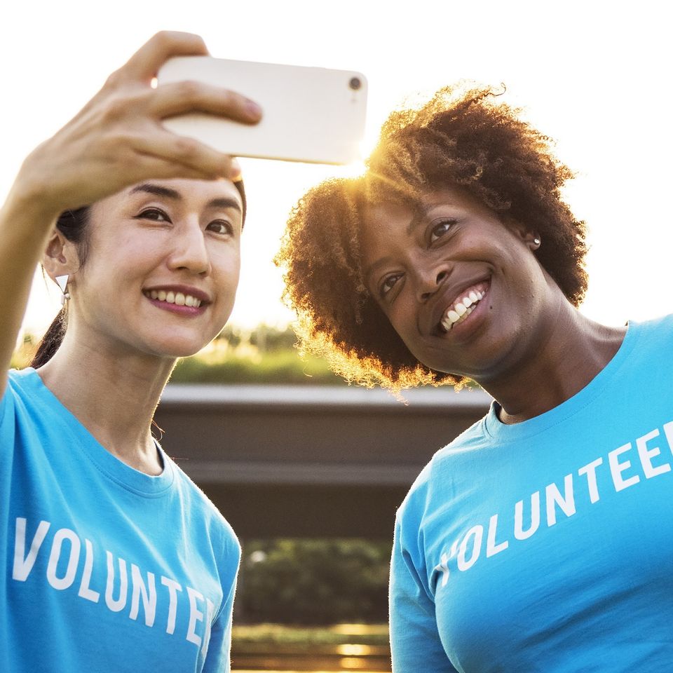 Two volunteers wearing matching blue volunteer shirts while taking a selfie outdoors during a community event.