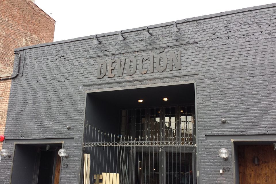 Grey dimensional letters reading Devoción mounted above a gated entrance on a painted dark grey brick facade