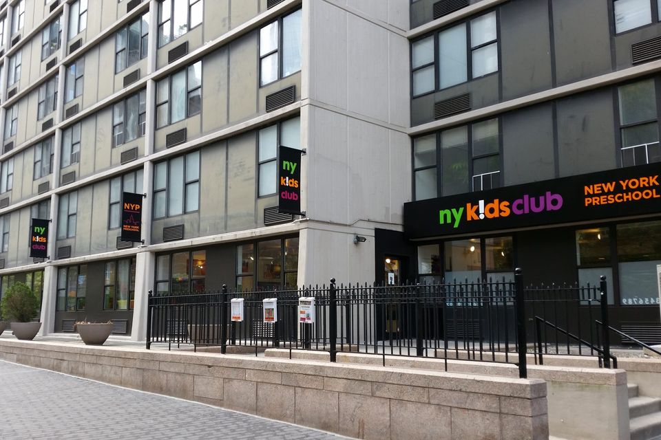 Row of NY Kids Club flag signs projecting along a building facade above a New York Preschool storefront sign in Battery Park