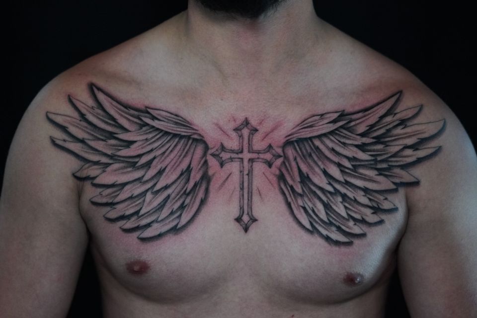 Image of a pair of wings tattood on a mans chest with a cross in the center between the wints