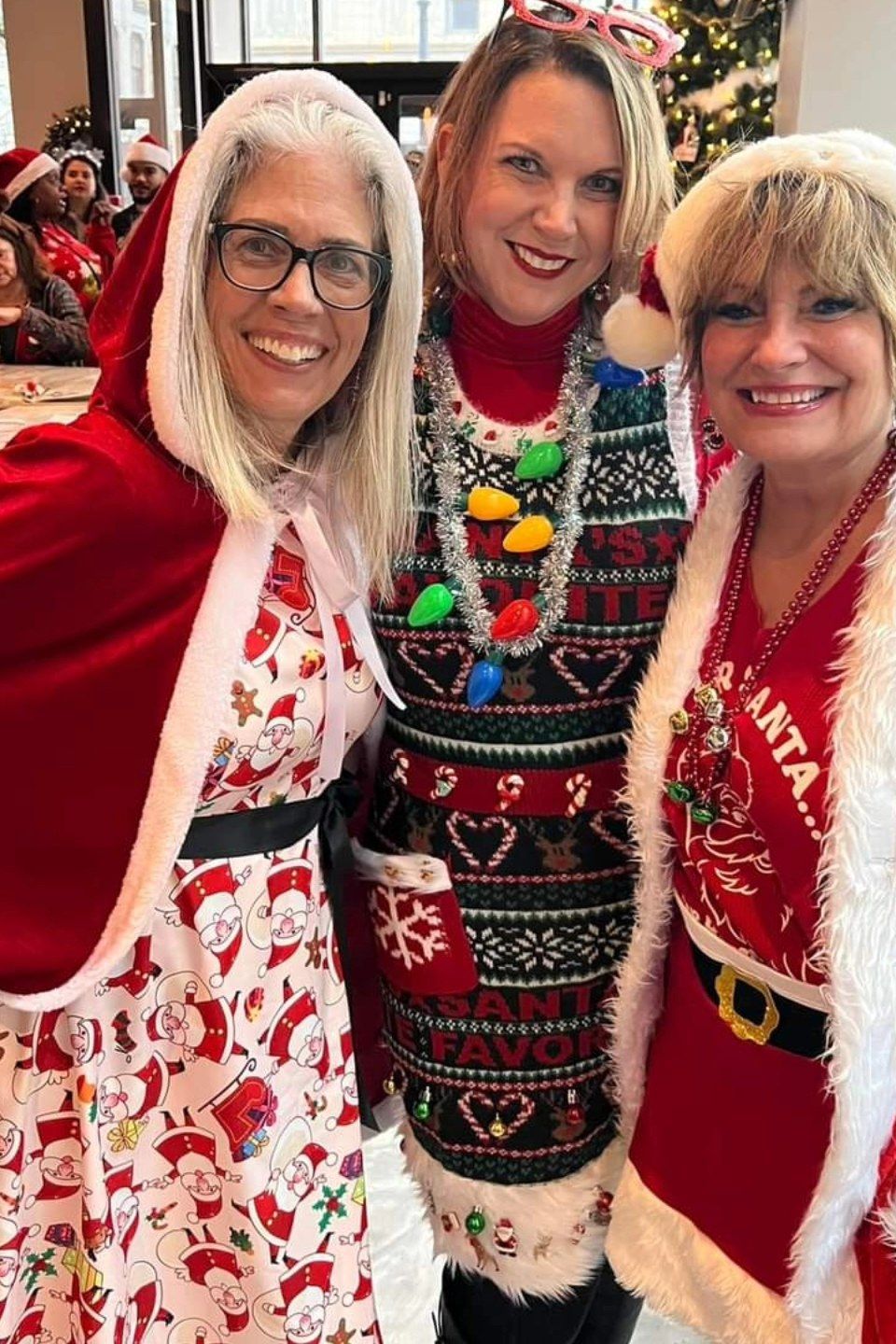 Three women wearing Christmas-themed clothing and accessories smiling for a photo indoors.