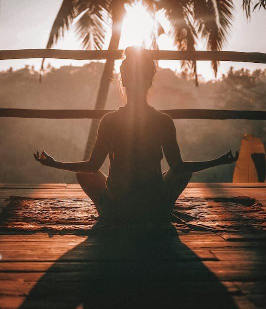 Person sitting on a deck in a meditative position with the sun beams shining through a palm tree 