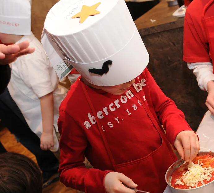 Child wearing a chef hat adding cheese to a personal pizza during a pizza-making activity.
