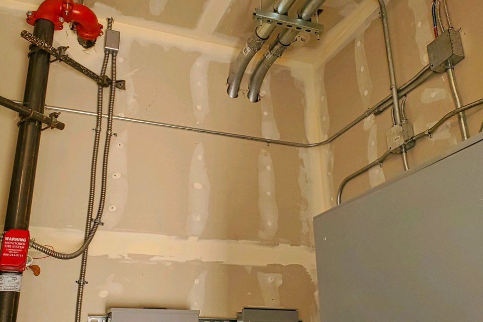 A technical view of a commercial electrical room featuring professionally bent metal conduit, junction boxes, and a fire system riser with red monitoring equipment.