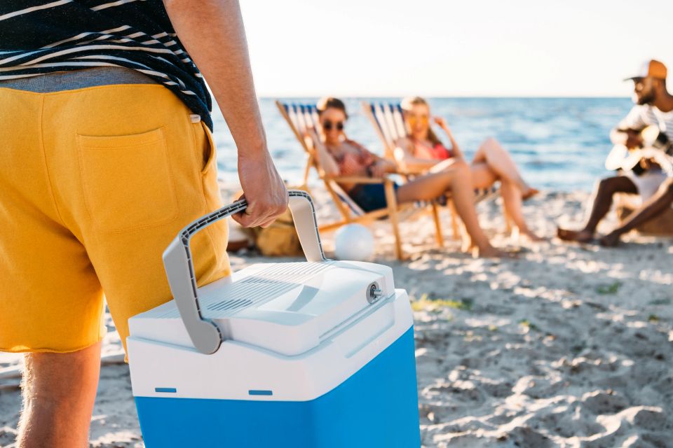 Man carrying a blue beach cooler toward friends relaxing in beach chairs on the sand