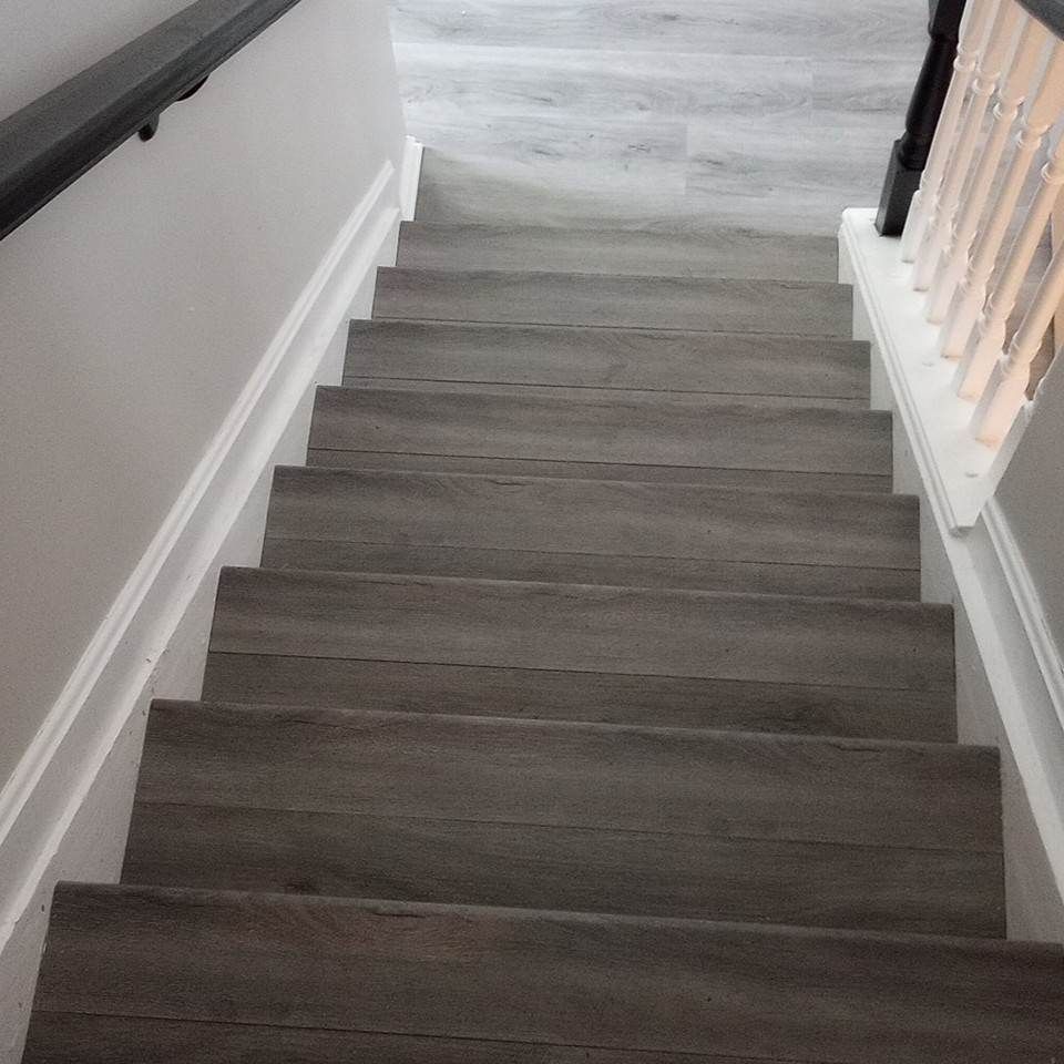 Gray wood-look flooring installed on a staircase with white railings and dark handrails.