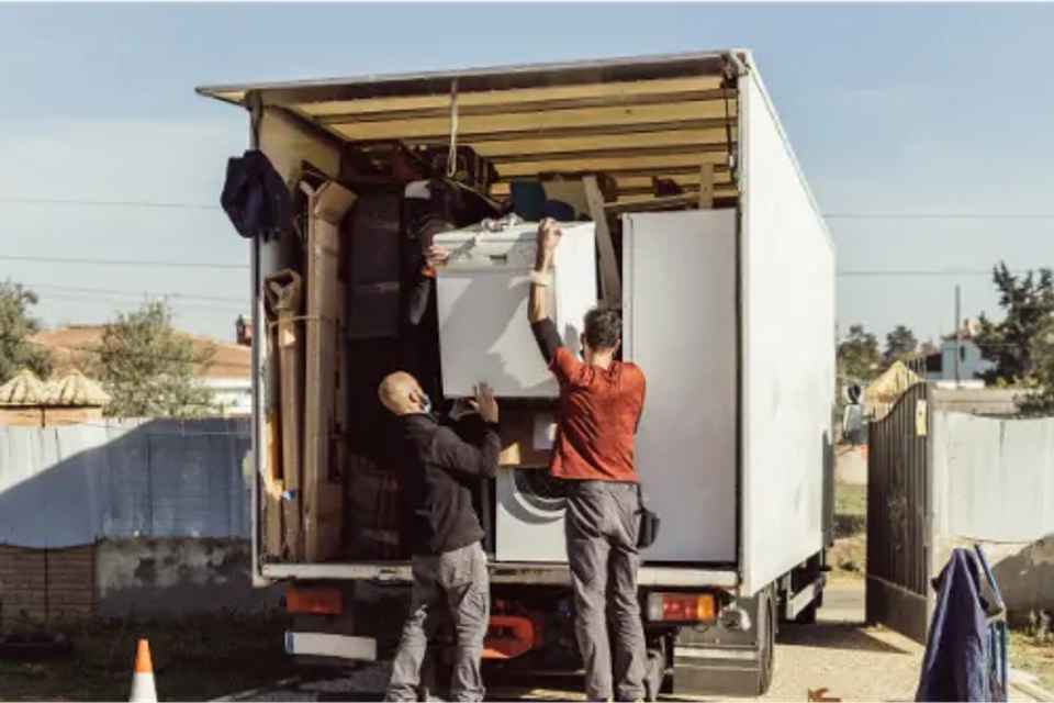 Two professional movers carefully lifting a heavy washing machine into a moving truck using proper lifting techniques.