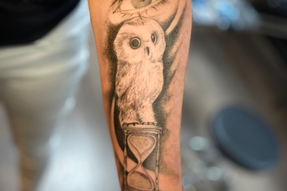 Image of a woman's forearm with a tattoo of X & O, an hour glass, a small owl, and an blue eye in black and grey and blue ink