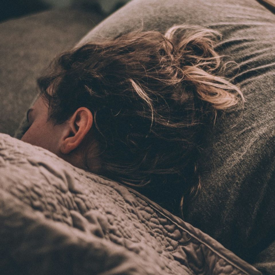 Woman sleeping in bed with part of her face visible under the covers, representing rest, fatigue, sleep quality, and low energy