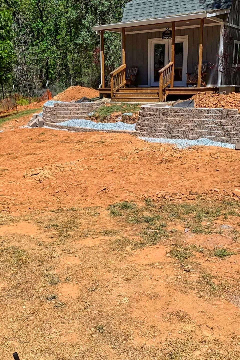 Pre-paving phase of a backyard renovation by Alta Vista Landscaping & Tree Service featuring a graded dirt site, newly constructed tiered block retaining walls, and a base layer of gravel for a custom patio installation near a garden cottage