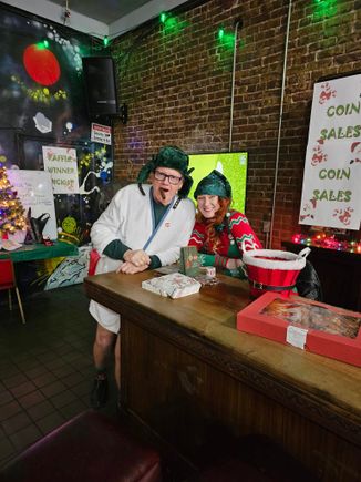Two people in festive holiday outfits standing behind a table at a Christmas-themed fundraiser.
