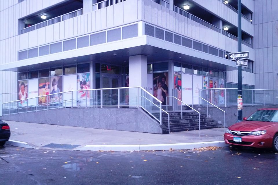 Printed window graphics with photos of children and NY Kids Club logos wrapping the ground floor windows of a corner building in Long Island City