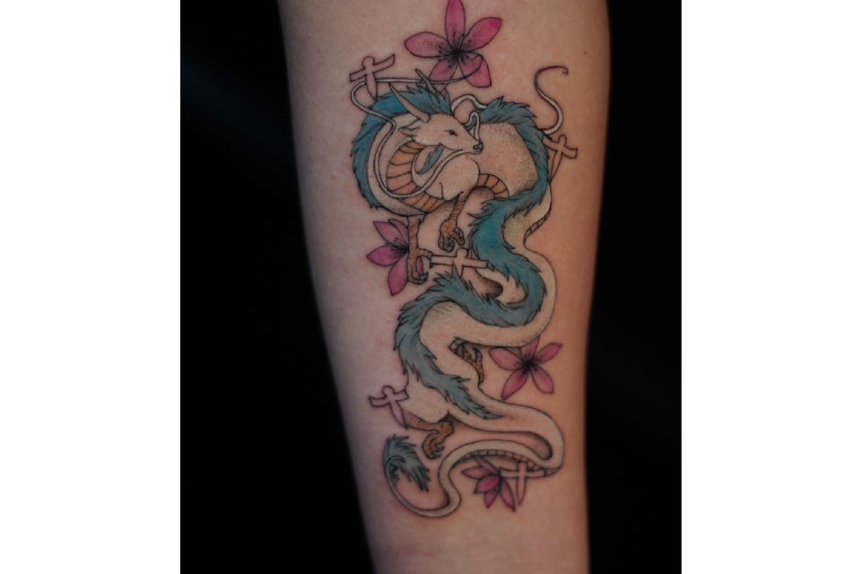 Image of a tattoo of a goat headed dragon in many colors, blue gold, with black outline and pink flowers and outlined crosses that are floating like angels around the dragon