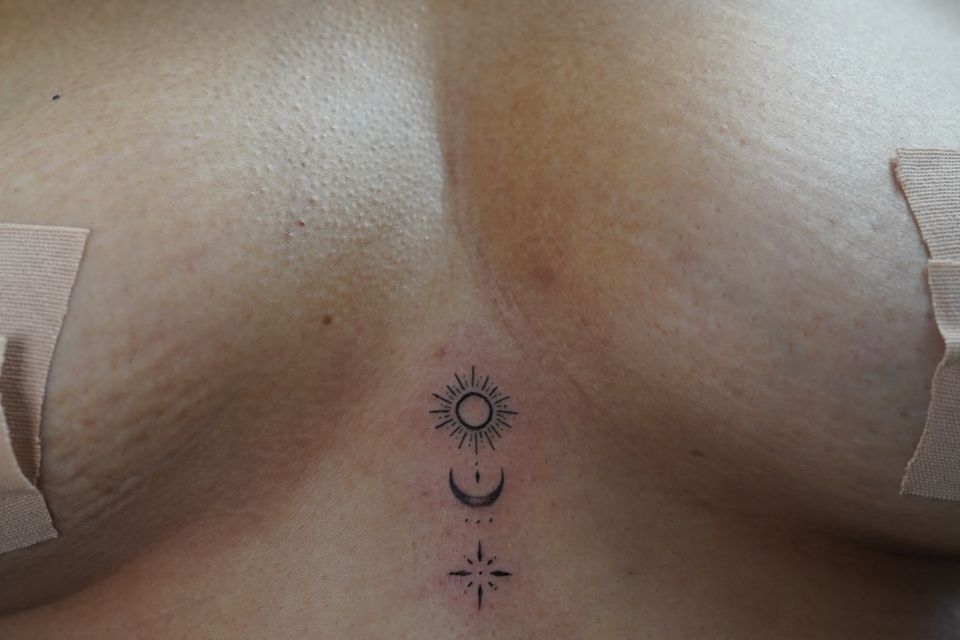 Image of a woman with a tattoo between her breasts with a small sun at the top, a crescent moon and star in the center and a star underneath it all in black and grey