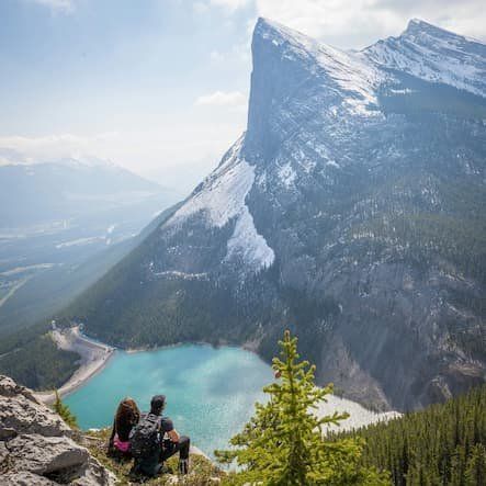 Two people sitting over a huge mountain visit with a lake below 