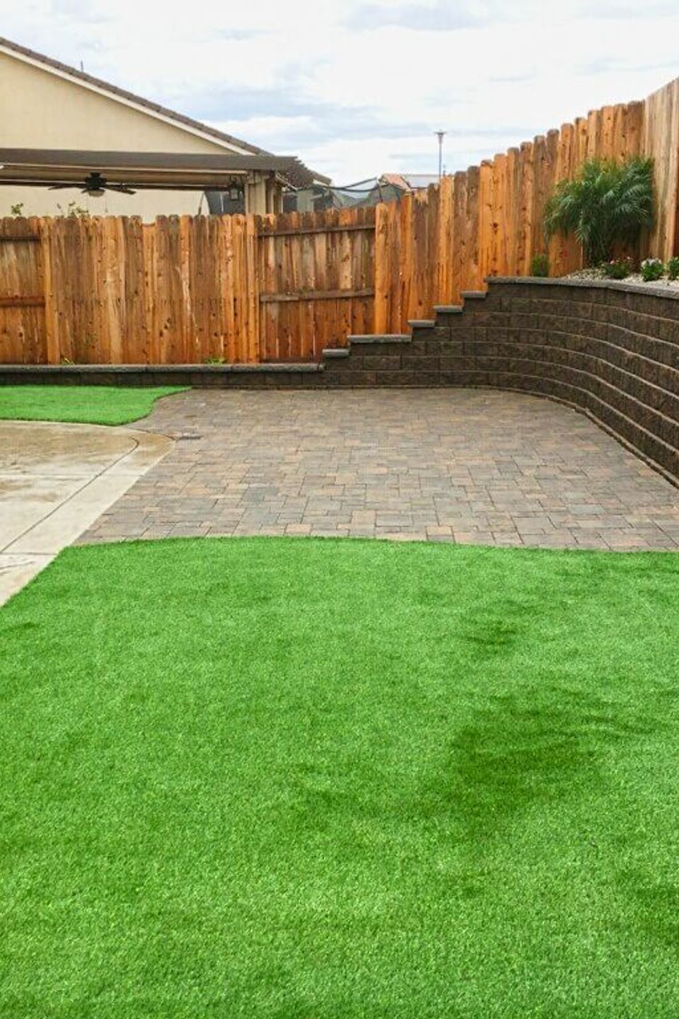 Modern low-maintenance backyard transformation featuring a tiered stone retaining wall, high-quality artificial turf, and a custom paver patio under a residential pergola.