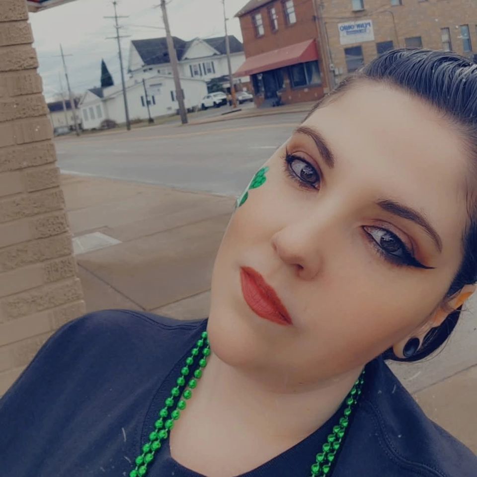Woman wearing green beads and shamrock face decoration posing for a selfie outdoors.