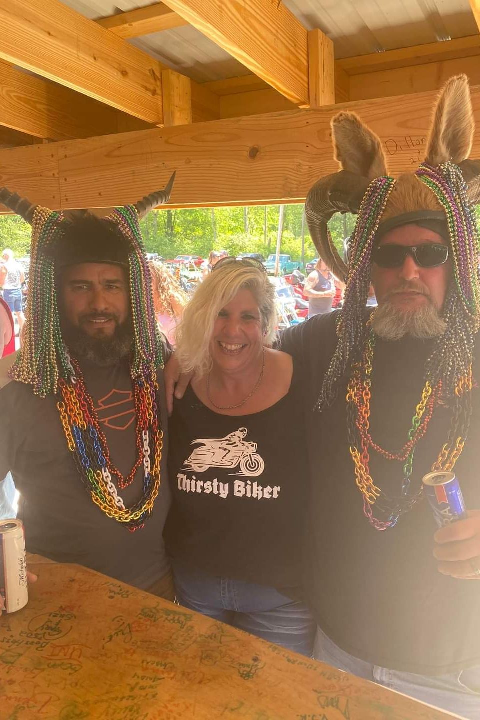 Three attendees posing together indoors, wearing horned headpieces decorated with colorful beads at a biker event.