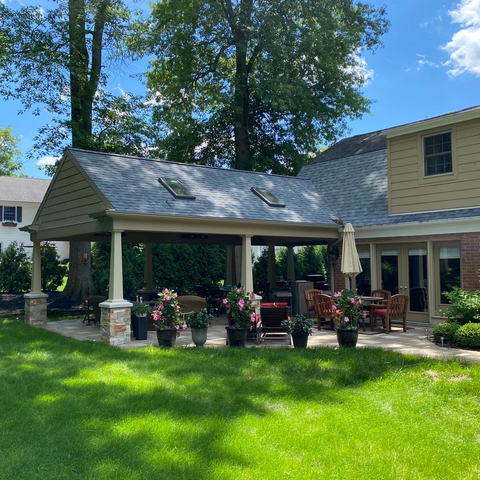 Covered backyard patio addition with stone columns, outdoor seating, dining area, and landscaped surroundings.