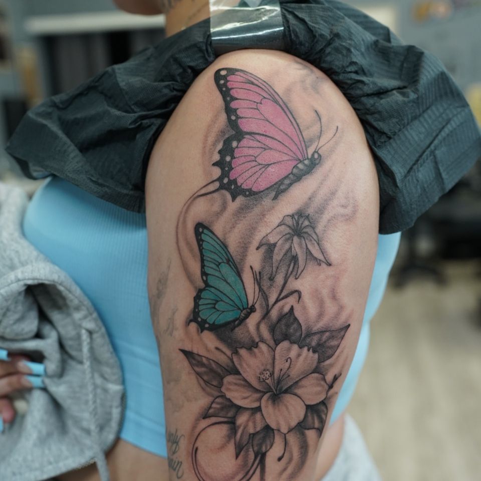 Image of a tattoo that includes 2 colorful butterflies, 1 pink and black and one teal and black, while the garden of flowers is in black and shades of grey