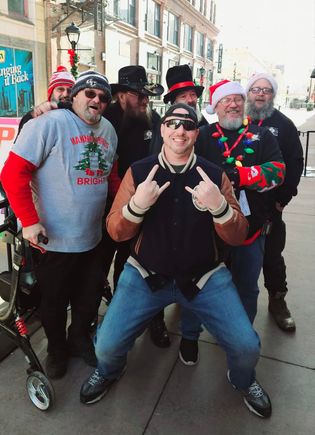 Group of attendees wearing Christmas hats, sweaters, and holiday accessories posing for a photo on a city sidewalk.