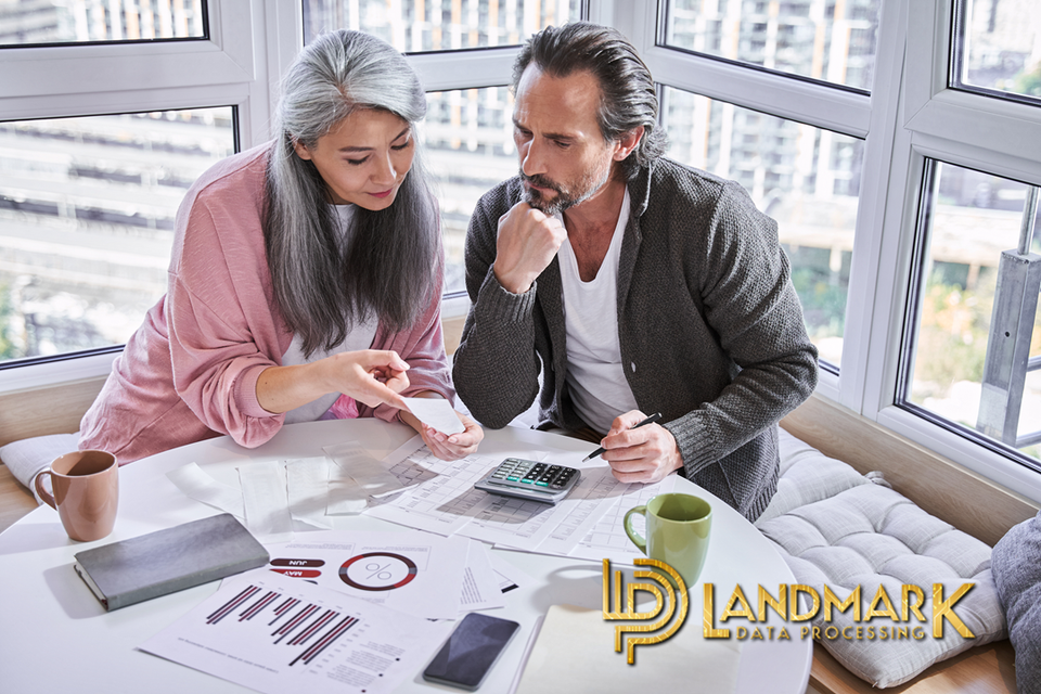 A woman and man at a table using a calculator to figure out why their payment processor is costing them so much. Choose Landmark Data Processing.