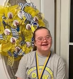 Smiling individual with Down syndrome wearing a DSAV shirt and medal while standing in front of a decorative blue and yellow wreath.