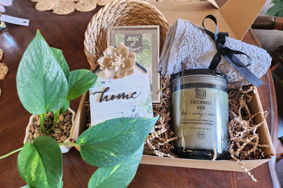 Gift box with potted plant, woven coaster, gray towel, patchouli candle, decorative cards, burlap flower, and brown crinkle paper on wooden table.” Home gift basket
