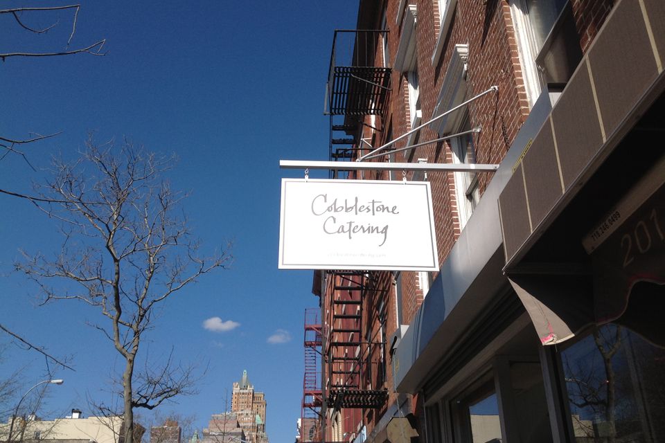 White hanging sign on chains from a wall bracket above a Brooklyn storefront against a clear blue sky