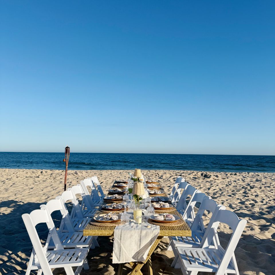 Long oceanfront dinner table with white folding chairs styled for a beach event in the Hamptons NY